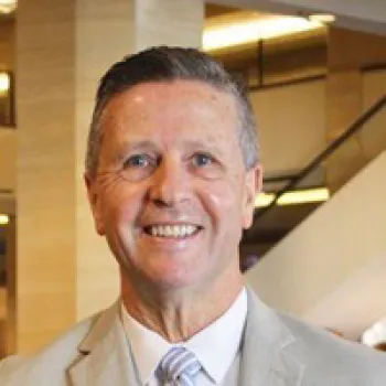 Shaun Robinson in a light-colored suit and striped tie smiles at the camera in an indoor setting with blurred background lights.
