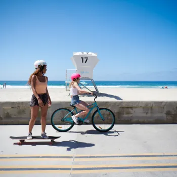 Two teenagers on a bike and skateboard cruising down the Board Walk of Mission Beach in San Diego