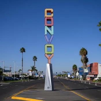A tall, colorful neon sign reading "CONVOY" stands on a median in the middle of a wide street lined with palm trees and businesses under a clear blue sky.
