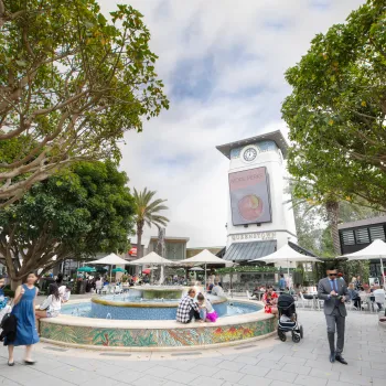 People walk and sit around a tiled fountain in an outdoor UTC Westfield plaza with trees, umbrellas, and a clock tower in the background.