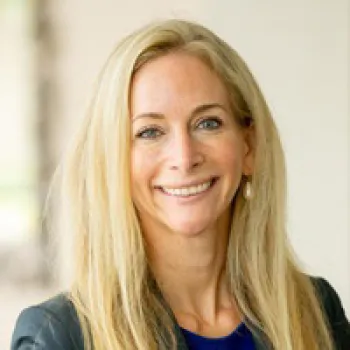 Lydia Bartell wearing a black blazer and blue top, smiles at the camera against a blurred light background.