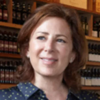 Tracy Borkum with shoulder-length brown hair stands in front of shelves stocked with bottles and wooden boxes, looking slightly to the side and smiling.