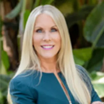 Ilsa Butler with long blonde hair wearing a teal blazer stands outdoors in front of green foliage, smiling at the camera.