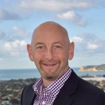 Daniel Kuperschmid wearing a checked shirt and dark blazer smiles in front of a coastal cityscape with a cloudy sky.