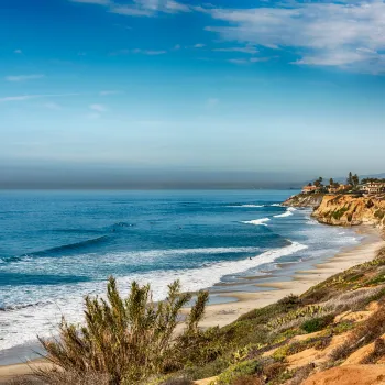 View of a sandy Carlsbad beach with waves rolling in, bordered by rocky cliffs and coastal vegetation under a partly cloudy sky.