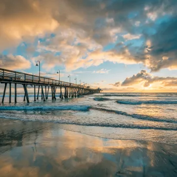 Imperial Beach pier stretches over the ocean under a dramatic sky at sunset, with waves washing onto the sandy beach and reflections on the wet sand.