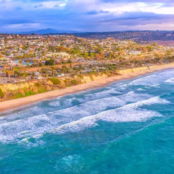 Aerial view of waves hitting Cardiff-by-the-Sea's sandy beach with a coastal town and hills in the background under a partly cloudy sky.