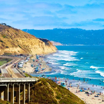 Crowded Torrey Pines State Beach along a coastline with cliffs, a road, and a railway; people are swimming, sunbathing, and walking near the waves under a partly cloudy sky.