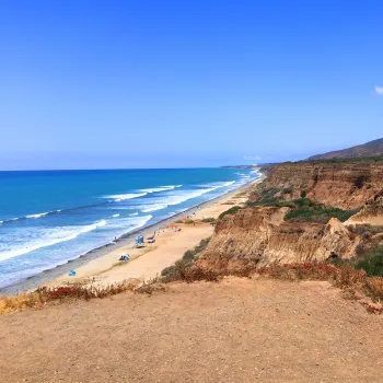 Coastal cliffs overlook the sandy San Onfre beach with scattered people and umbrellas; waves approach shore under a clear blue sky with distant hills in the background.