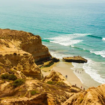 Cliffs with a winding staircase overlook a sandy Blacks Beach beach and ocean waves, with people walking along the shore and a rocky outcrop.
