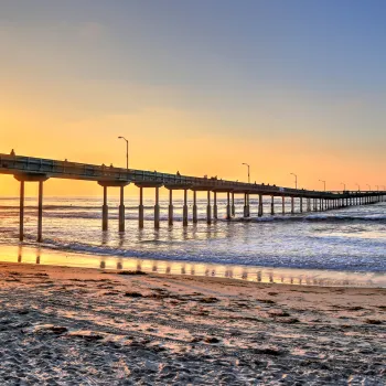 Ocean Beach pier extends over the ocean at sunset, with a surfer carrying a board on the sandy beach in the foreground.
