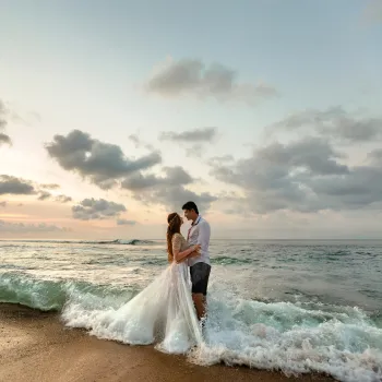 A bride and groom stand together at the shoreline, embracing in the surf at sunset with clouds scattered across the sky.