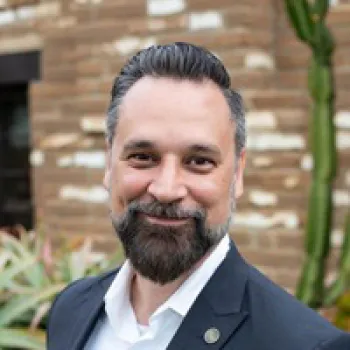 Eric Jenkins with dark hair and beard wearing a suit jacket and white shirt, smiling outdoors in front of a brick wall and plants.