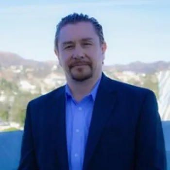 Jamie Lemon with light skin, short brown hair, and a beard is wearing a dark suit and blue shirt, standing outdoors with a blurred cityscape and hills in the background.
