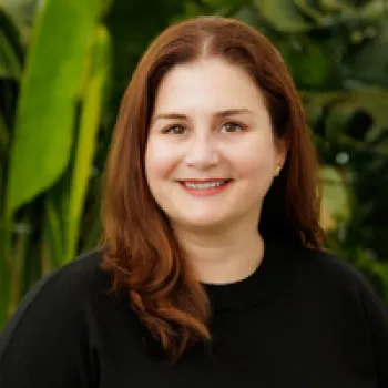 Maria Valdes with long brown hair wearing a black top smiles at the camera, standing in front of greenery.