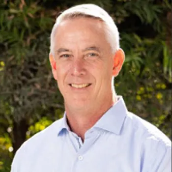 Todd McAninch with short gray hair wearing a light blue button-up shirt, standing outdoors with greenery in the background.