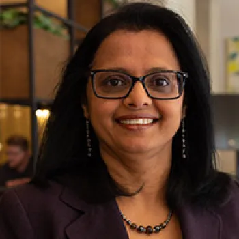 Cecilia Menezes with straight black hair, wearing glasses, a dark blazer, earrings, and a beaded necklace, smiling at the camera in an indoor office setting.