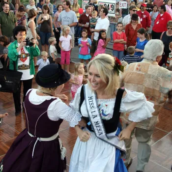 People in traditional clothing dance at an outdoor Oktoberfest in San Diego while a crowd of onlookers, including children, watch. A woman wearing a "Miss La Mesa" sash is smiling in the center.