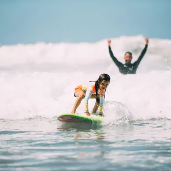 A young girl begins to stand up on a surfboard in the whitewater of a wave on a sunny day at a San Diego Beach, as her instructor raises his arms joyously behind her.