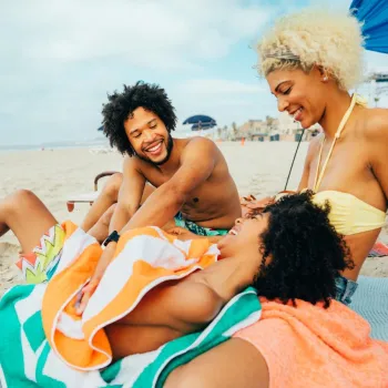 Three people sit smiling and laughing together on a beach, sitting on towels under a canopy with sand and ocean visible in the background.