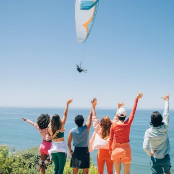 Six people stand on a grassy bluff at the Torrey Pines Gliderport in the La Jolla neighborhood of San Diego, facing the ocean with arms raised as they watch a paraglider flying over the water under a clear blue sky.