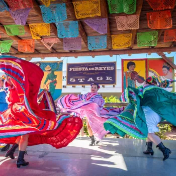 Three dancers in colorful traditional dresses perform on an outdoor stage at Fiesta de Reyes in San Diego's Old Town, decorated with papel picado banners and Fiesta de Reyes signs in the background.