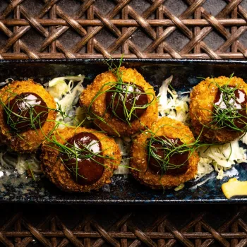 Six breaded, fried croquettes topped with sauce and herbs are arranged on shredded cabbage in a rectangular blue plate at Realm of the 52 Remedies in San Diego.