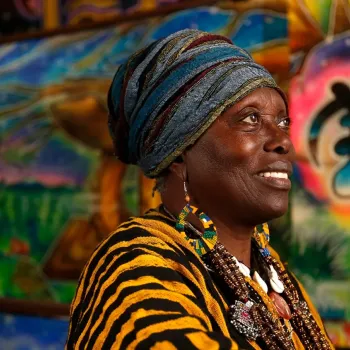 A woman in a colorful headwrap and patterned clothing stands in front of a vibrant, multi-colored mural, smiling and looking to the side.