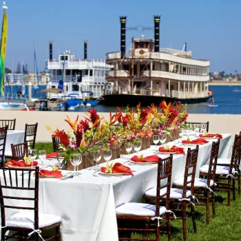 Two long tables with white tablecloths and floral centerpieces are set up on grass by the water, with sailboats and a large riverboat in the background.