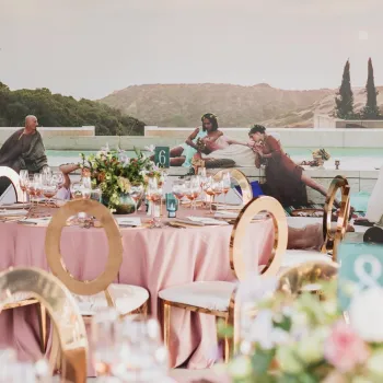 Outdoor event with a round table set for dining in the foreground; people in costumes recline on steps by a pool in the background with greenery and hills visible.