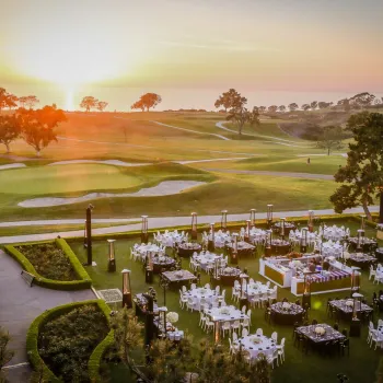 Outdoor event setup with round tables and chairs arranged on a lawn next to a golf course at sunset, with trees and a glowing sky in the background.