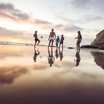 A group of people stand and play with a ball on a wet sandy beach at sunset, with cliffs and ocean visible in the background.