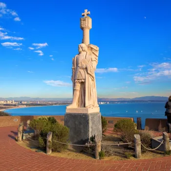 A stone statue of a historical figure stands on a raised platform overlooking the ocean, with people observing the coastal view and city skyline in the background.