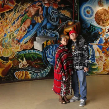 Two people stand closely together in front of a large, colorful mural featuring abstract and cultural imagery.