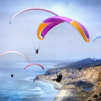 Several paragliders fly above coastal cliffs and the ocean under a cloudy sky, with a sandy shoreline and hilly landscape in the background.