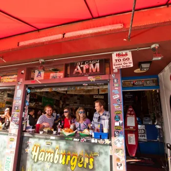 People sit at a counter eating food at an outdoor hamburger restaurant decorated with various signs and license plates under a red awning.