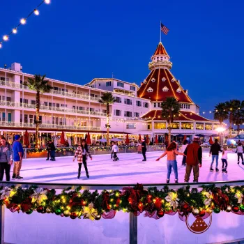 People ice skating on a decorated outdoor rink at dusk, with a large historic hotel and festive holiday lights in the background.