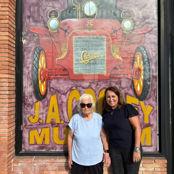 Two women stand smiling in front of a mural of a red antique car outside the J.A. Cooley Museum, which specializes in antique cars.