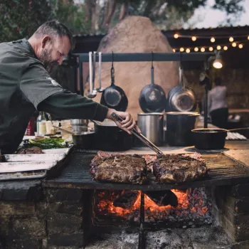 A man in a chef’s coat grills large pieces of meat over an open flame in an outdoor kitchen with hanging pots and string lights.