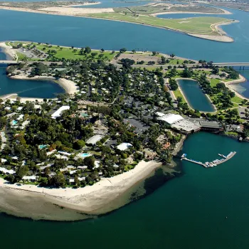 Aerial view of a small, lush island connected by a bridge, surrounded by water with sandy shores and greenery, featuring buildings and a dock.