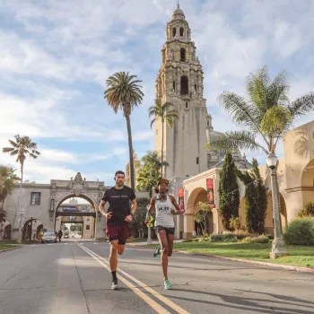 Two people run down a street lined with palm trees and historic buildings, with a tall tower in the background under a partly cloudy sky.
