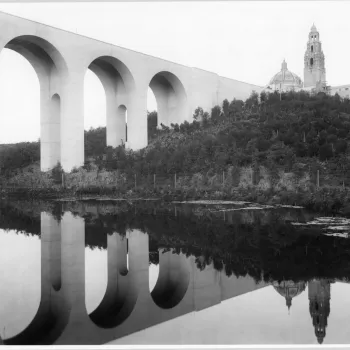 A large arched bridge spans over a river, reflecting in the water below, with a domed building and tower visible on a wooded hill in the background.