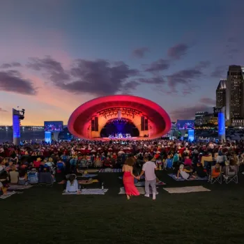 The Shell. A large crowd sits on a lawn facing an outdoor concert stage with a red-lit arch, city skyscrapers, and a sunset sky in the background.