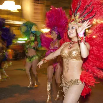 People in colorful feathered costumes and headdresses parade down a street at night, with blurred lights and motion in the background.