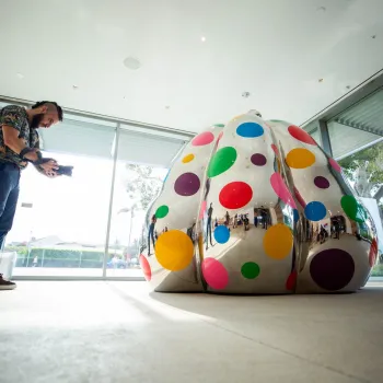 A man photographs a large, reflective sculpture covered in colorful polka dots inside a modern building with glass doors and windows.