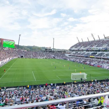 A large soccer stadium filled with spectators shows a match in progress on a green field under a partly cloudy sky.
