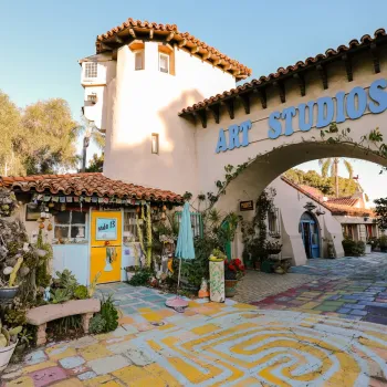 Colorful outdoor art studio with painted walkway, potted plants, tiled roofs, and a large “ART STUDIOS” sign above an archway on a sunny day.