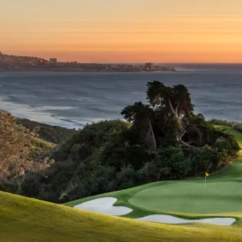 Torrey Pines North Golf Course 15 green with sand bunkers overlooks the ocean at sunset, with distant hills and a coastal town visible in the background.