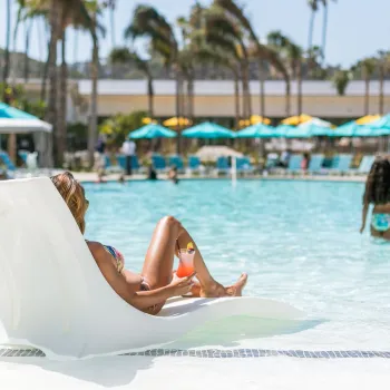 A person lounges on a pool chair in shallow water with a drink, while two children play nearby. Palm trees, umbrellas, and resort buildings are visible in the background.