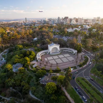 Aerial view of Balboa Park’s Spreckels Organ Pavilion in San Diego, with city skyline and a plane flying overhead in the background.